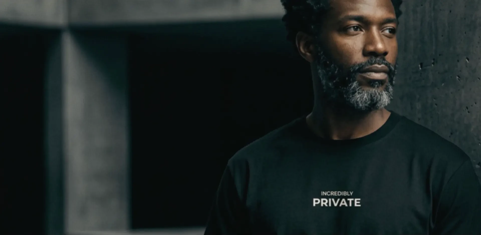 Man in a black T-shirt printed INCREDIBLY PRIVATE, standing beside concrete architecture in low light
