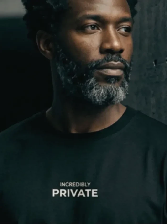 Man in a black T-shirt printed INCREDIBLY PRIVATE, beside concrete architecture in low light